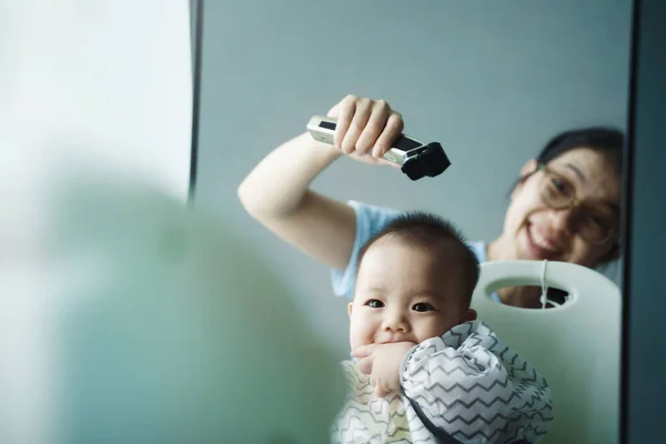 Cheerful Chinese mother playing with little son while he is getting haircut at home