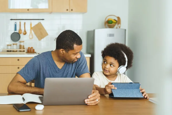 Black African American father working on laptop computer while his Afro Little son using digital tablet in kitchen at home.