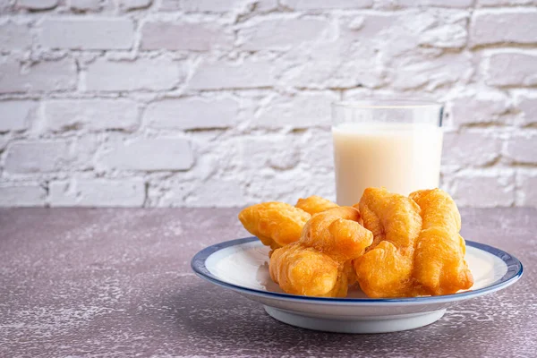 Close-up of deep-fried dough sticks or Chinese doughnut sticks on white plate and a glass of soybean milk on gray stone background with white brick wall. Space for text