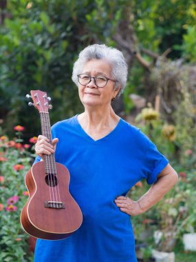 Bahçede dururken elinde ukulele tutan yaşlı bir Asyalı kadının portresi. Şarkı söyleyerek, küçük gitar çalarak, mutlu bir şekilde ve emekli olduktan sonra hayatın tadını çıkararak. Yaşlılar ve sağlık hizmetleri kavramı.