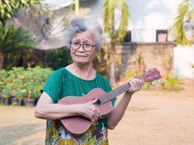 Bahçede dururken elinde ukulele tutan yaşlı bir Asyalı kadının portresi. Şarkı söyleyerek, küçük gitar çalarak, mutlu bir şekilde ve emekli olduktan sonra hayatın tadını çıkararak. Yaşlılar ve sağlık hizmetleri kavramı.