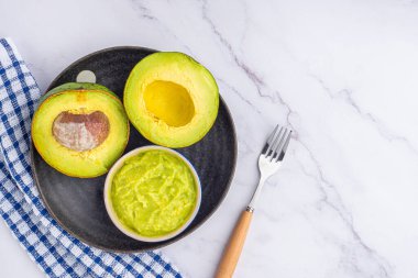 Tohumlu organik avokado, avokado yarımları, ve siyah tabakta taze guacamole mermer zemin üzerinde kumaş ve çatal bıçak. Üst Manzara. Düz yatış stili. Sağlıklı meyve kavramı..