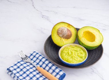 Tohumlu organik avokado, avokado yarımları, ve siyah tabakta taze guacamole mermer zemin üzerinde kumaş ve çatal bıçak. Üst Manzara. Düz yatış stili. Sağlıklı meyve kavramı..