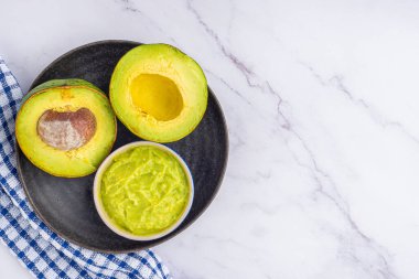Organic avocado with seed, avocado halves, and fresh guacamole in a bowl on a black plate with cloth on a marble table. Top view. Flat lay style.Concept of healthy fruit.