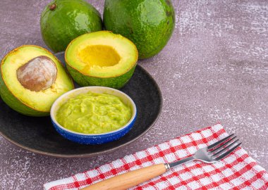 Organic avocado with seed, avocado halves, and fresh guacamole in a bowl on a black plate with cloth and cutlery on a gray stone background. Top view. Space for text. Concept of healthy fruit.