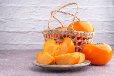 Fresh ripe persimmons with slices of persimmon on a plate and whole fruit in a basket with a white brick wall. Space for text. Close-up photo. Concept of health fruit.
