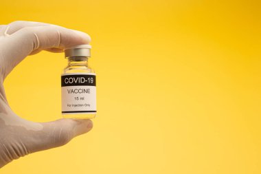 Elinde COVID-19 aşı şişesi ve sarı arka planı olan hijyenik eldiven giyen bir elin yakın çekimi. Coronavirus aşısı. Mesaj için yer var. Tıp ve sağlık hizmetleri kavramı.