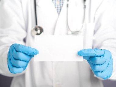 Hastanede dikilirken beyaz bir kağıt tutan koruyucu eldivenler giymiş bir doktorun ellerinin yakın çekimi. Mesaj için yer var. Tıbbi ve iletişim konsepti.