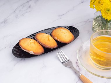 Siyah bir tabakta, mermer bir fonda beyaz tuğla duvarlı bir fincan çayla limon jöleli kurabiyeler. Madeleine - ev yapımı geleneksel Fransız küçük kurabiyesi. Deniz kabuğu tatlı kekleri.