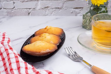 Siyah bir tabakta, mermer bir fonda beyaz tuğla duvarlı bir fincan çayla limon jöleli kurabiyeler. Madeleine - ev yapımı geleneksel Fransız küçük kurabiyesi. Deniz kabuğu tatlı kekleri.