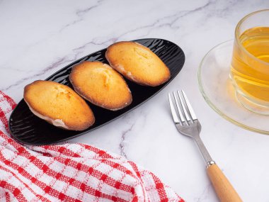 Siyah bir tabakta, mermer bir fonda beyaz tuğla duvarlı bir fincan çayla limon jöleli kurabiyeler. Madeleine - ev yapımı geleneksel Fransız küçük kurabiyesi. Deniz kabuğu tatlı kekleri.