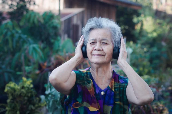 Kablosuz kulaklık takan son sınıf Asyalı kadın bahçede dikilirken en sevdiği şarkıyı dinliyor. Yaşlılar ve rahatlama kavramı.