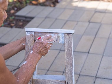 Senior man holding a brush to paint white on the old wooden chair without a backrest. Old chairs are painted white. Renovation of old furniture.