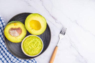 Tohumlu organik avokado, avokado yarımları ve siyah bir tabakta mermer zemin üzerinde kumaş ve çatal bıçakla taze guacamole. Üst Manzara. Düz yatış stili. Sağlıklı meyve kavramı..