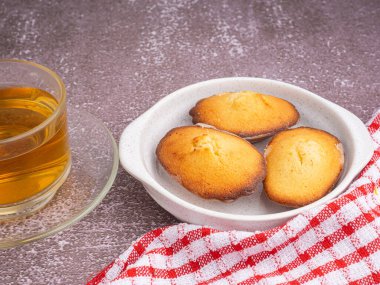 Siyah bir tabakta limon jöleli kurabiyeler ve gri taşlı arka planda bir fincan çay. Madeleine - ev yapımı geleneksel Fransız küçük kurabiyesi. Mesaj için yer var. Deniz kabuğu tatlı kekleri.