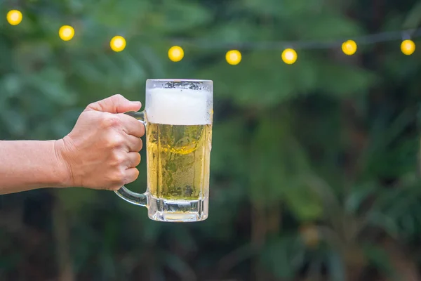 Hand holding a mug of beer at a party in the garden background. Close-up photo. Space for text. Concept of party and beverages.