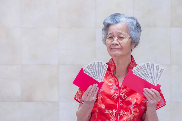 Elderly Asian woman wearing a Cheongsam red shirt, holding a red envelope packet with US dollars (or Ang Pao) for giving to children during Chinese New Year. Space for text. Concept of festival.