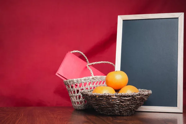 A small blackboard, four tangerines, and red envelopes (Ang Pao) in a basket placed on a wooden table with a red cloth background. Space for text. Happy Chinese New Year concept.