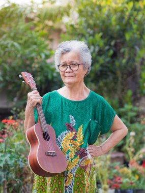 Cheerful elderly Asian woman with short gray hair, wearing glasses, holding the ukulele, and looking at the camera while standing in the garden. Space for text. Concept of aged people and relaxation.