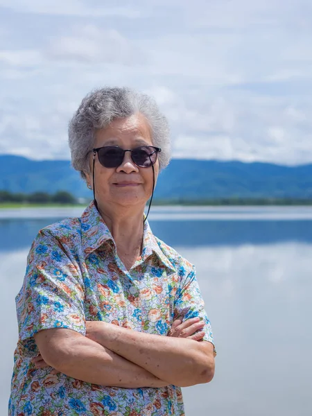 Portrait of senior woman wearing sunglasses arms crossed, smiling and looking at the camera while standing beside the lake with mountains backgrounds. Concept of Aged people and relaxation.