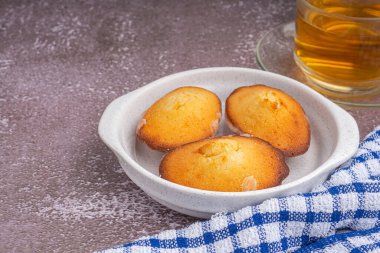 Lemon glaze madeleine on white a dish with a teacup placed on a gray stone background. Madeleine - homemade traditional French small cookie. Space for text. Seashell sweet cakes.
