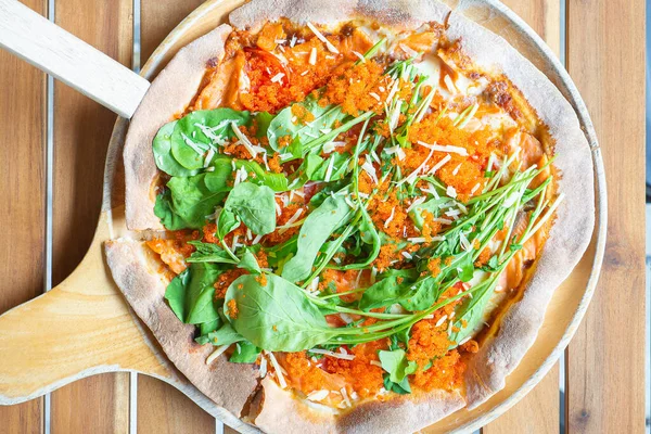 Pizza salmon and rocket consisted of tomato sauce, mozzarella cheese, smoked salmon, rocket, tomato, ebiko, parmesan cheese on a wooden plate placed on a wood table. Concept of healthy food.