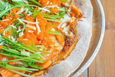 Pizza salmon and rocket consisted of tomato sauce, mozzarella cheese, smoked salmon, rocket, tomato, ebiko, parmesan cheese on a wooden plate placed on a wood table. Concept of healthy foods.