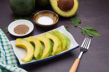 The sliced avocados are placed on a white plate with cutlery and cloth placed on a dark gray background. Space for text. Close-up photo. Concept of healthy fruits.