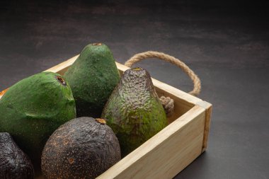 Avocados are placed in a wooden crate on a dark gray background. Space for text. Close-up photo. Concept of healthy fruits.
