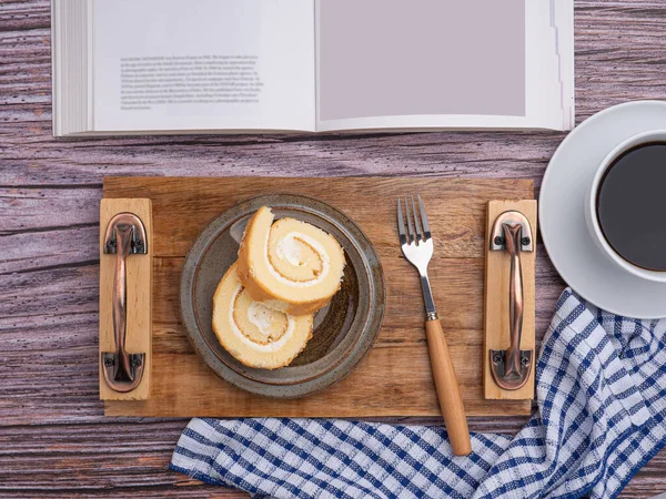 Top view of a book with roll cake on a plate on a wooden tray and a white coffee cup placed on a wooden table. Space for text. Concept of relaxation.