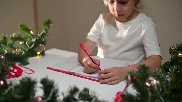 身穿白色T恤 头戴红笔 头戴新年帽的女孩在桌上给亲爱的圣诞老人写了一封信 希望他早产 愿新年礼物的清单 — 图库视频影像