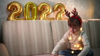 Noel geyiği bantlı mutlu bir kız hediye kutusunu açar. Arka planda 2022 altın sayılı mutlu noeller konsepti var.