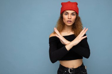 Şaşkın, hoş, çekici, esmer, samimi duyguları olan, siyah kırbaç bluzu giyen, mavi arka planda izole edilmiş kırmızı şapkalı ve dur hareketi yapan bir kadın. Olumsuz