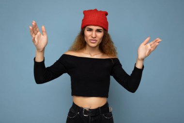 Genç, duygusal tatminsiz, güzel esmer, dalgalı saçlı, samimi duyguları olan, siyah bluz giyen ve mavi arka planda izole edilmiş kırmızı şapkalı bir kadın.