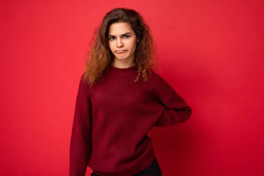 Genç, tatminsiz, üzgün, kıvırcık esmer, samimi duyguları olan, arka plandaki duvarda izole edilmiş, koyu kırmızı süveter giyen bir kadın. Bozuk konsept.