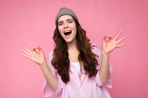 Портрет молодой позитивной счастливой эмоционально привлекательной курчавой брюнетки с искренними эмоциями в стильной розовой рубашке и серой шляпе, изолированной на розовом фоне с копировальным пространством и показывающей в порядке — стоковое фото
