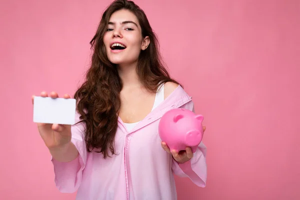 Hermosa feliz alegre joven morena con camisa aislada sobre fondo rosa con espacio libre y sosteniendo caja de dinero de cerdo rosa y tarjeta de crédito para maqueta — Foto de Stock