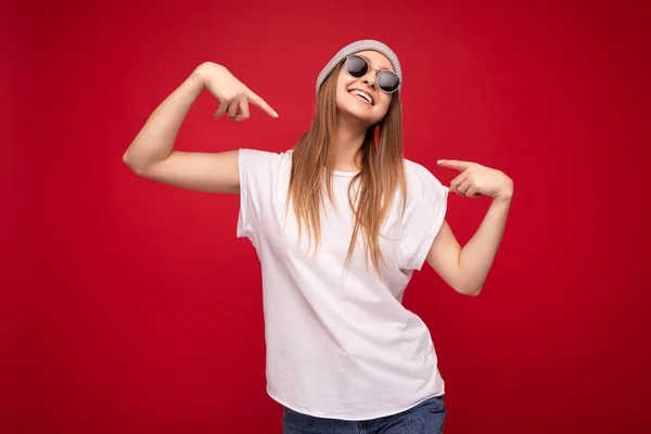 Foto de una joven mujer rubia atractiva atractiva y positiva con emociones sinceras usando una camiseta blanca casual con espacio vacío para sombrero gris y gafas de sol aisladas sobre fondo rojo —  Fotos de Stock