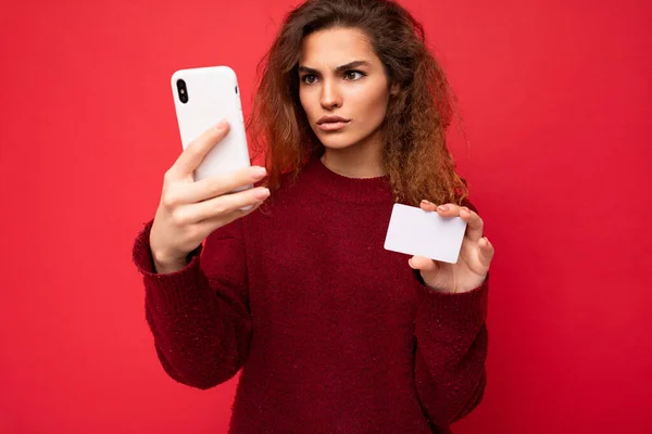Young dissatisfied beautiful curly brunette woman with sincere emotions poising isolated over background wall with empty space wearing casual dark red sweater holding credit card and using mobile — Stock Photo, Image