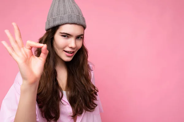 Portrét mladé pozitivní šťastný okouzlující kudrnaté brunetky žena s upřímnými emocemi na sobě stylové růžové košile a šedý klobouk izolované na růžovém pozadí s kopírovacím prostorem a ukazuje ok gesto — Stock fotografie