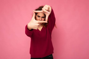 Samimi duyguları olan genç, pozitif esmer, kıvırcık kadın portresi. Pembe arka planda izole edilmiş, fotokopi çekilen pembe kazak giyiyor ve el kamerasıyla fotoğraf çeken el hareketlerini gösteriyor.