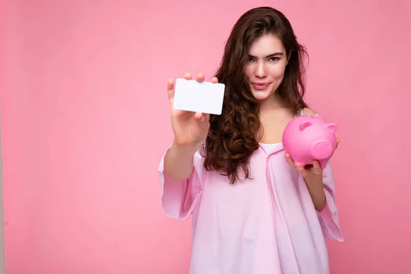 Portrét krásné pozitivní veselý roztomilý usmívající se mladá bruneta žena ve stylové košili izolované na růžovém pozadí s kopírovacím prostorem a držení růžové prasečí penízky a kreditní karty pro mockup — Stock fotografie
