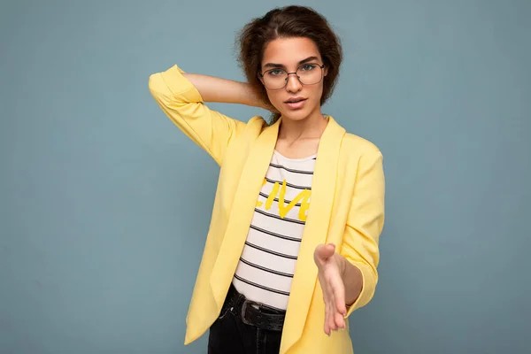 Foto de una joven morena atractiva y segura de sí misma con emociones sinceras usando una elegante chaqueta amarilla y gafas ópticas aisladas sobre fondo azul con espacio para copiar — Foto de Stock