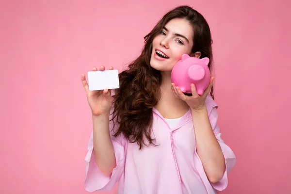 Portrét krásné pozitivní veselý roztomilý usmívající se mladá bruneta žena ve stylové košili izolované na růžovém pozadí s kopírovacím prostorem a držení růžové prasečí penízky a kreditní karty pro mockup — Stock fotografie