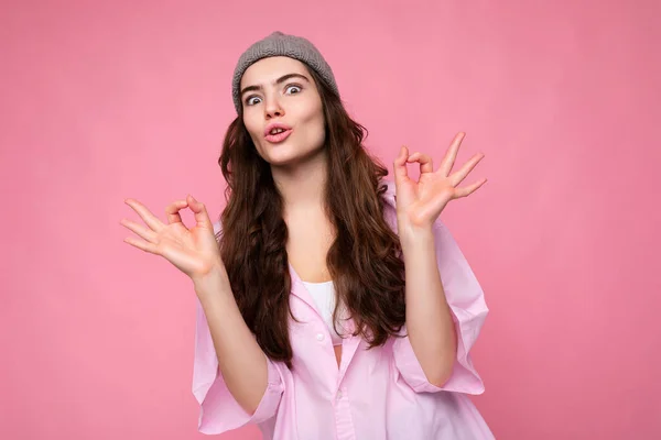 Portrait of young positive surprised beautiful curly brunette woman with sincere emotions wearing trendy pink shirt and grey hat isolated on pink background with empty space and showing ok gesture. It — Stock Photo, Image