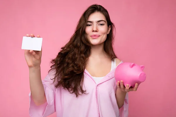 Atraktivní šťastná mladá brunetka žena nosí košili izolované na růžovém pozadí s prázdným prostorem a drží růžové prase peníze box a kreditní karty pro mockup — Stock fotografie