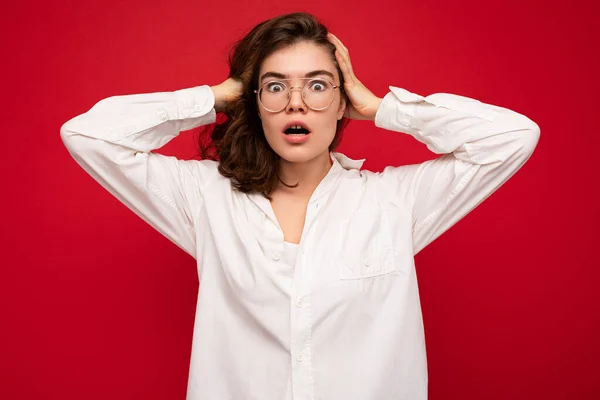 Attraente giovane emotivo scioccato stupito donna bruna riccia indossa camicia bianca e occhiali ottici isolati su sfondo rosso con spazio copia — Foto Stock