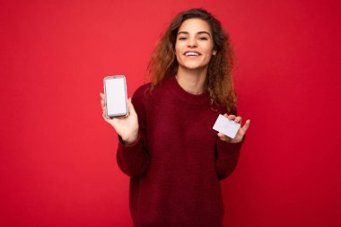 Samimi duyguları olan genç, kıvırcık esmer bir kadın arka plan duvarında yalnız başına kalmış, boş bir yerde, koyu kırmızı süveter giymiş, elinde kredi kartı cep telefonu olan, boş ekranlı bir cep telefonu tutan.
