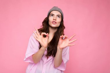 Genç, duygusal, komik, çekici, kıvırcık esmer kadının portresi. Samimi duyguları olan, şık pembe gömlekli, pembe arka planda izole edilmiş gri şapkalı, fotokopi odası olan ve iyi olduğunu gösteren bir kadın.