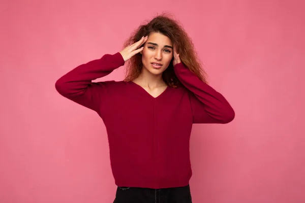 Portrait of young emotional beautiful brunette curly woman with sincere emotions wearing trendy pink pullover isolated on pink background with copy space and having headache — Stock Photo, Image